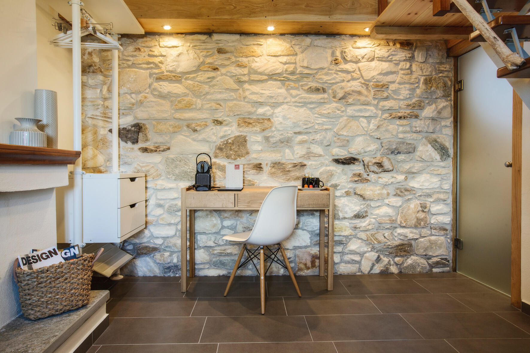 Πέτρινος τοίχος με ξύλινο γραφείο, λευκή μοντέρνα καρέκλα και διακριτικό φωτισμό, σε γωνιά δωματίου boutique ξενοδοχείου. Stone wall with wooden desk, modern white chair and soft lighting in a cozy corner of a boutique hotel room.