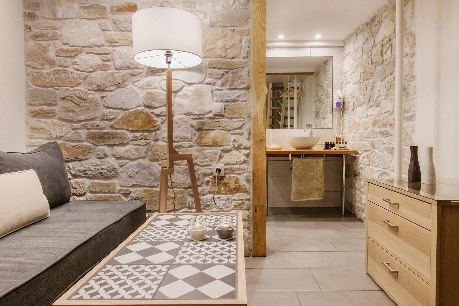 Μια κομψή γωνιά με φυσική πέτρα και ξύλο, όπου το design συναντά τη λειτουργικότητα σε ένα δωμάτιο boutique ξενοδοχείου. Stone wall with wooden desk, modern white chair and soft lighting in a cozy corner of a boutique hotel room.