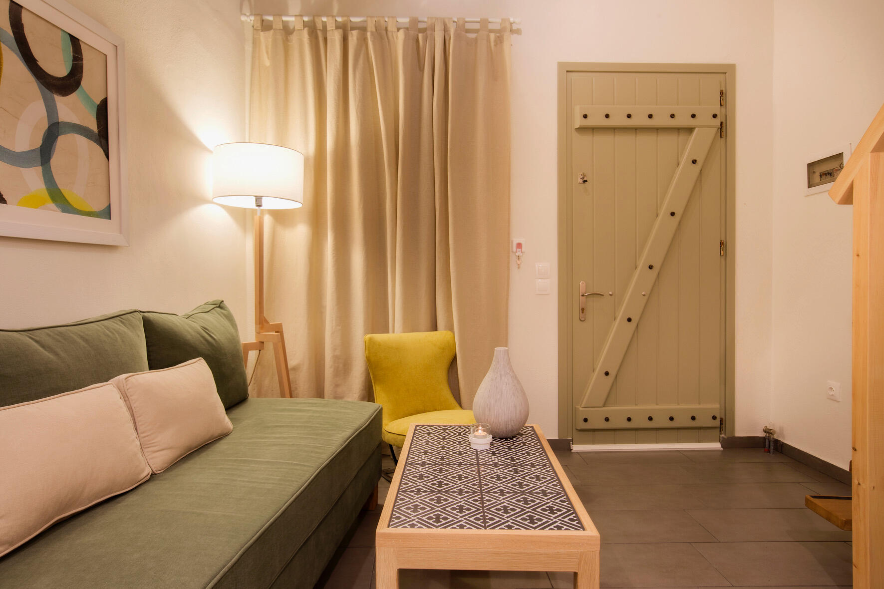 Καθιστικός χώρος σε δωμάτιο ξενοδοχείου με πράσινο καναπέ, κίτρινη μοντέρνα πολυθρόνα και custom τραπεζάκι από οξειά και πλακίδια ρετρό, με μπεζ κουρτίνες και minimal αισθητική. Seating area in a hotel room featuring a green sofa, a modern yellow armchair,